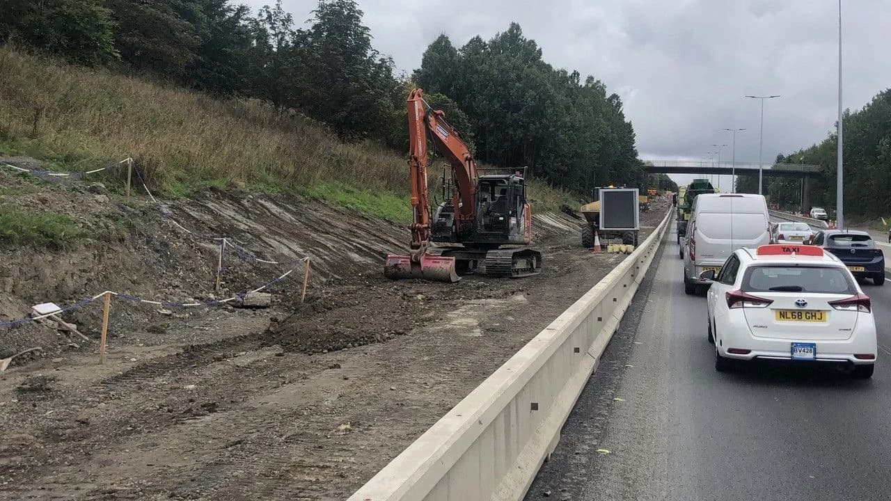 An orange excavator digs beside a motorway at Woolsington where a concrete barrier separates lane-restricted traffic queuing past the roadworks; no people