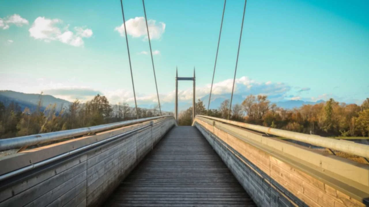 Wooden pedestrian suspension footbridge crossing a valley, with autumn trees and mountains in the distance.