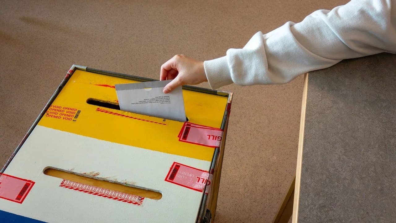 A hand dropping a ballot into a sealed yellow ballot box; no faces visible.