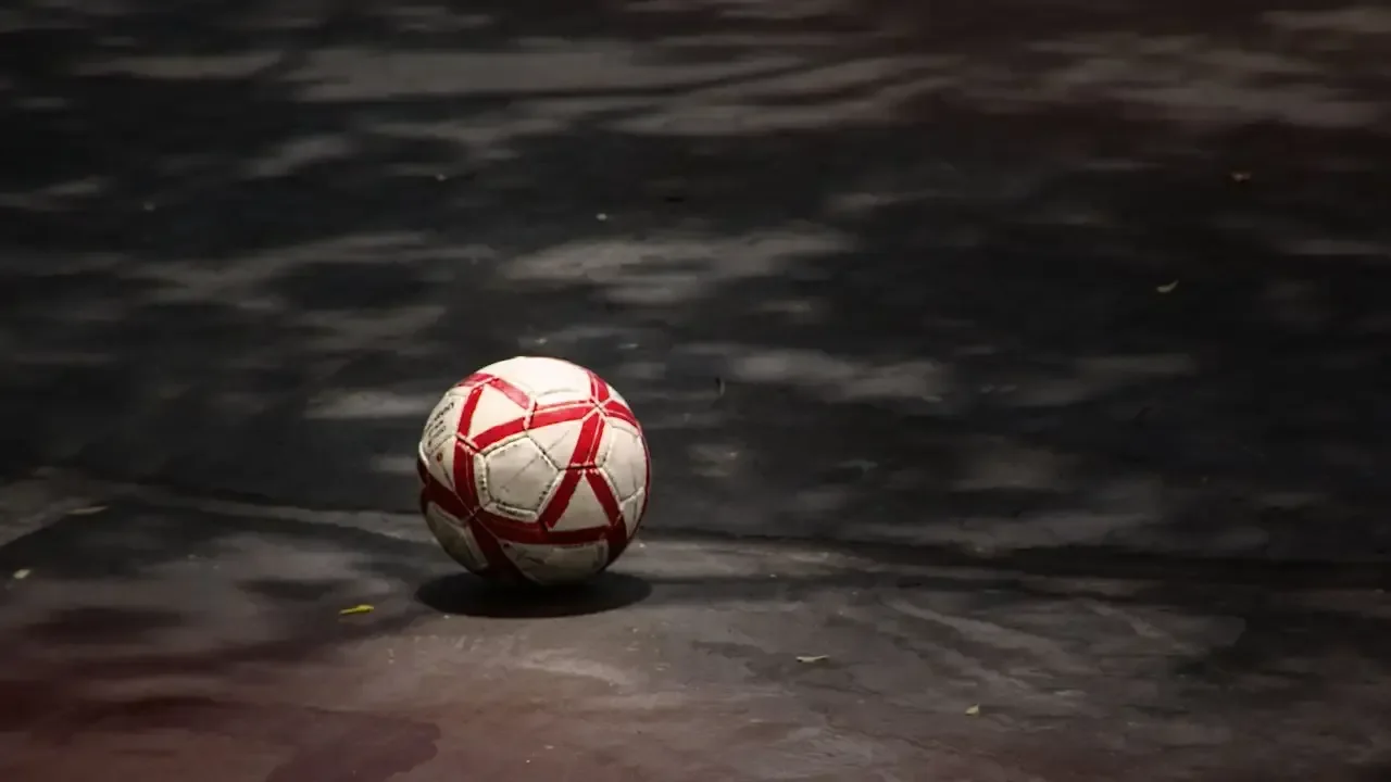 A red-and-white soccer ball resting alone on dark pavement dappled with tree shadows.